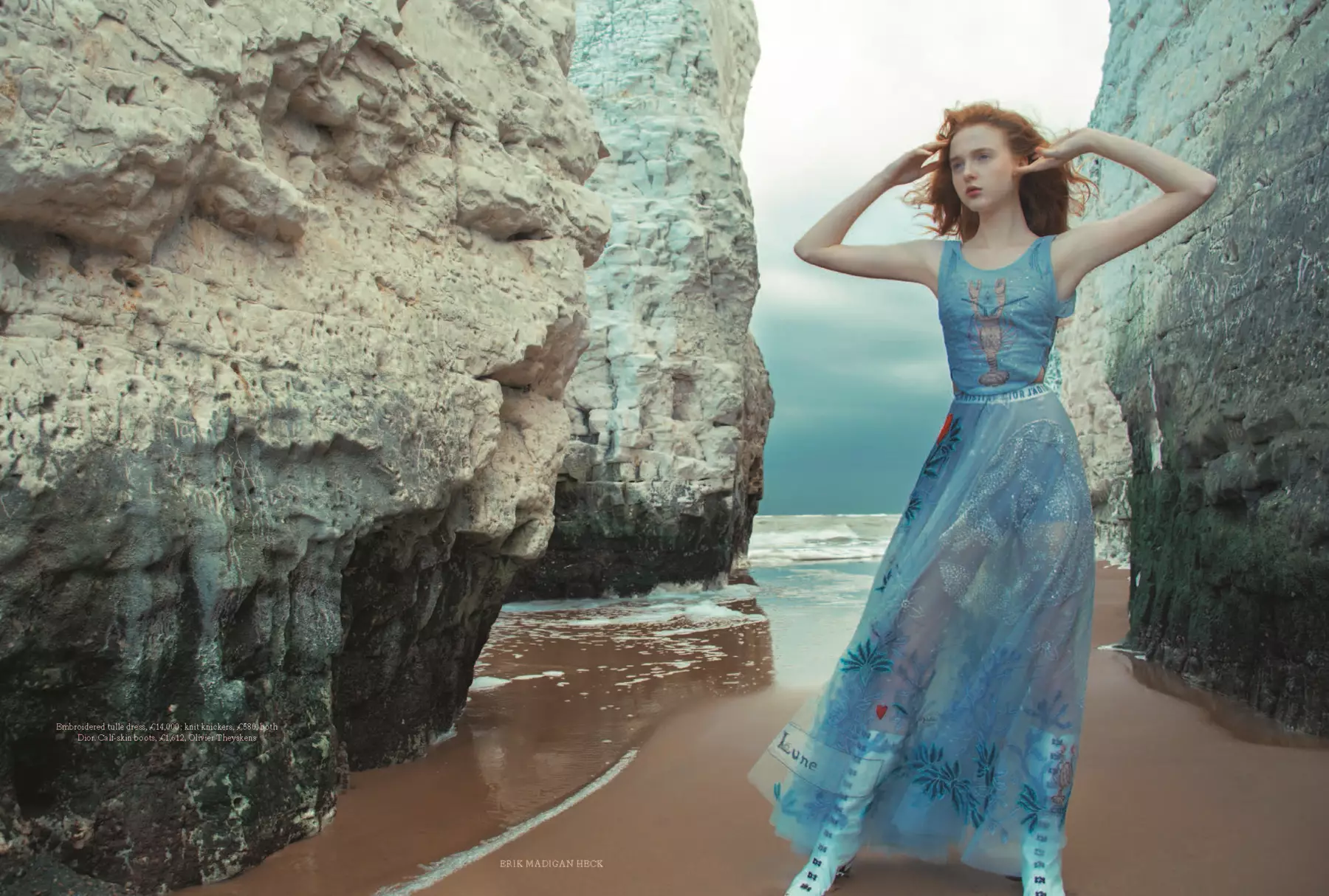 Lady in blue flowing dress with lobster logo on top half, stood between two chalk stacks on the beach. Bit of sea around feet and dark blue moody sky